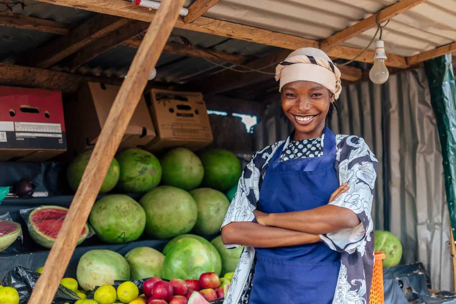 attractive African sales woman on blue apron isolated over fruit stand with her arms folded while looking at camera