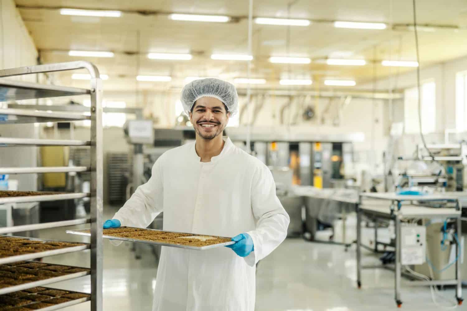 Cheerful multicultural food factory worker carrying fresh baked biscuits on tray.