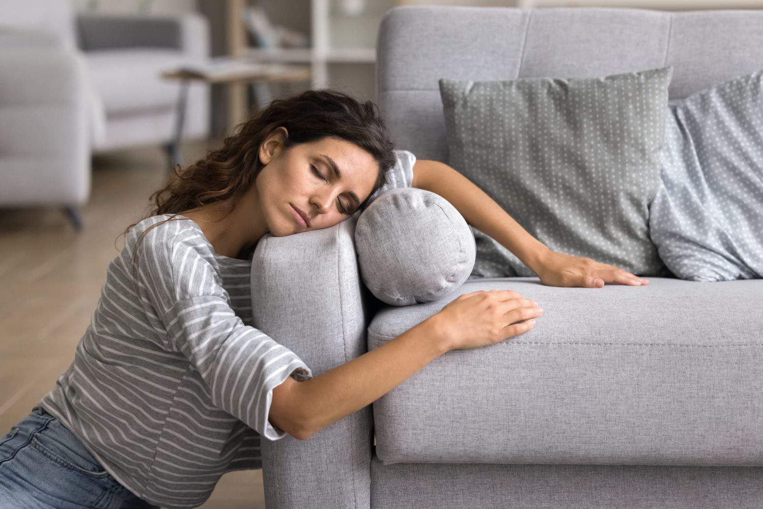 Exhausted sleepy young woman resting on floor at home, leaning head and hands on grey soft couch, enjoying leisure, break with closed eyes, accidently falling asleep near sofa