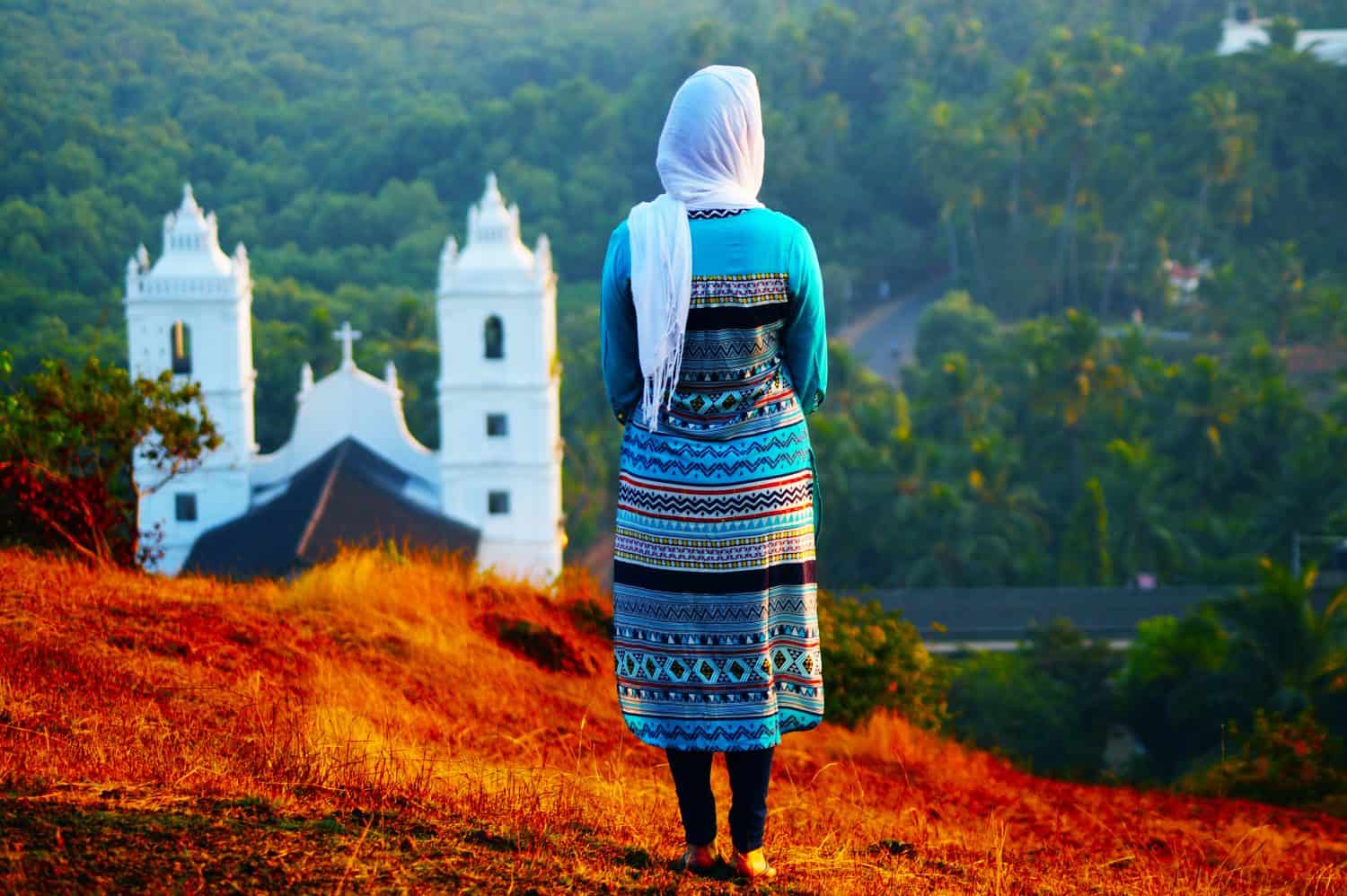 Young woman on the background of church. Her head covered with a scarf. She&#039;s in traditional Indian clothes. Sunset time. Moment of the religious life. Christian stand on the hill near the church.