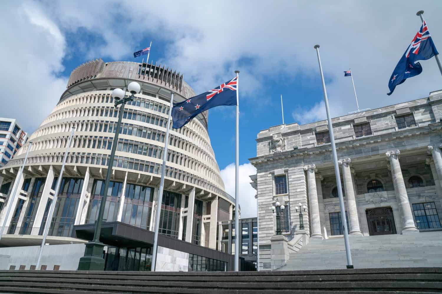 Flags flutter in front New Zealand Government buildings, House neo classical style House of Parliament with Beehive behind.