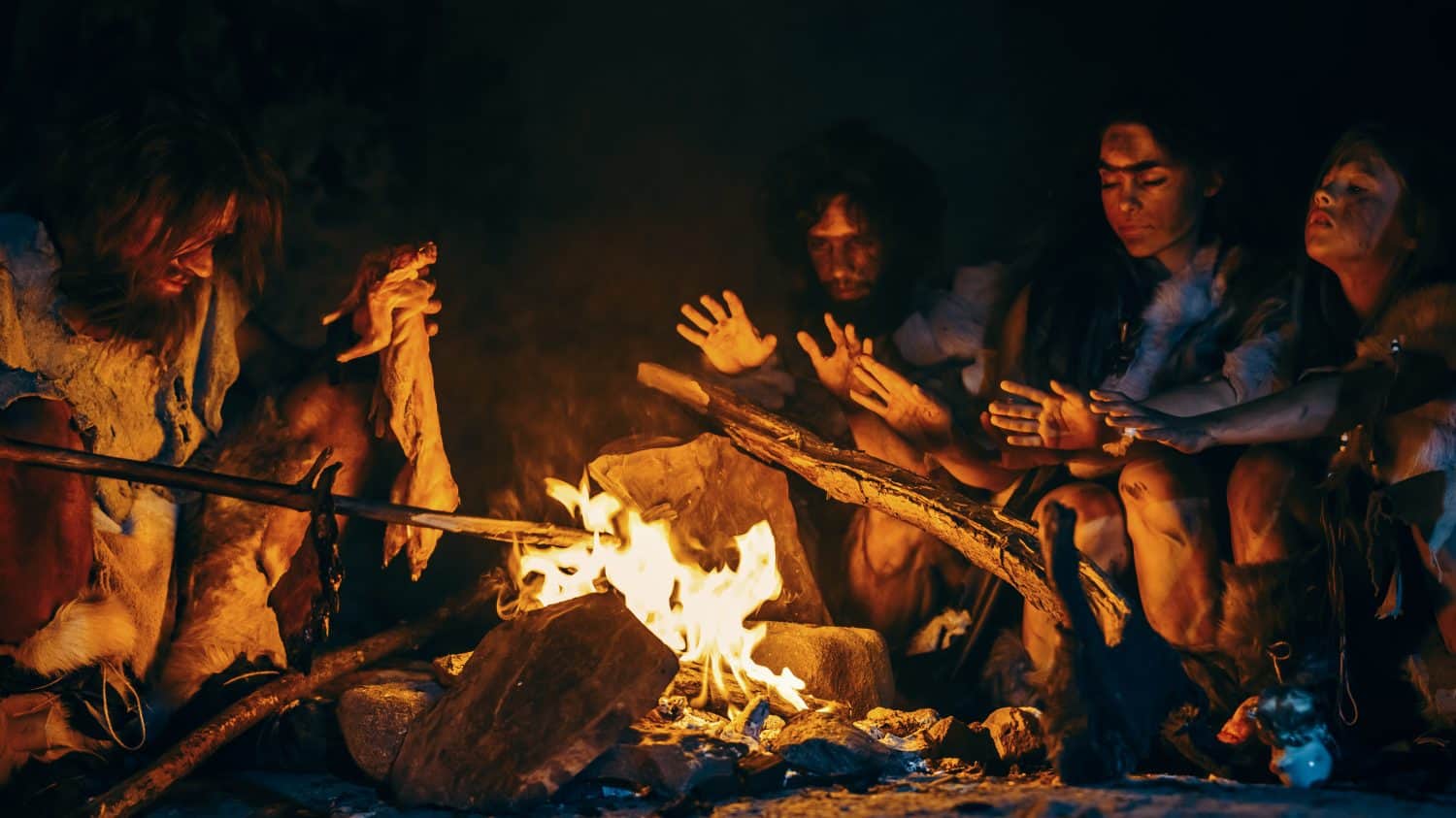 Neanderthal or Homo Sapiens Family Cooking Animal Meat over Bonfire and then Eating it. Tribe of Prehistoric Hunter-Gatherers Wearing Animal Skins Eating in a Dark Scary Cave at Night