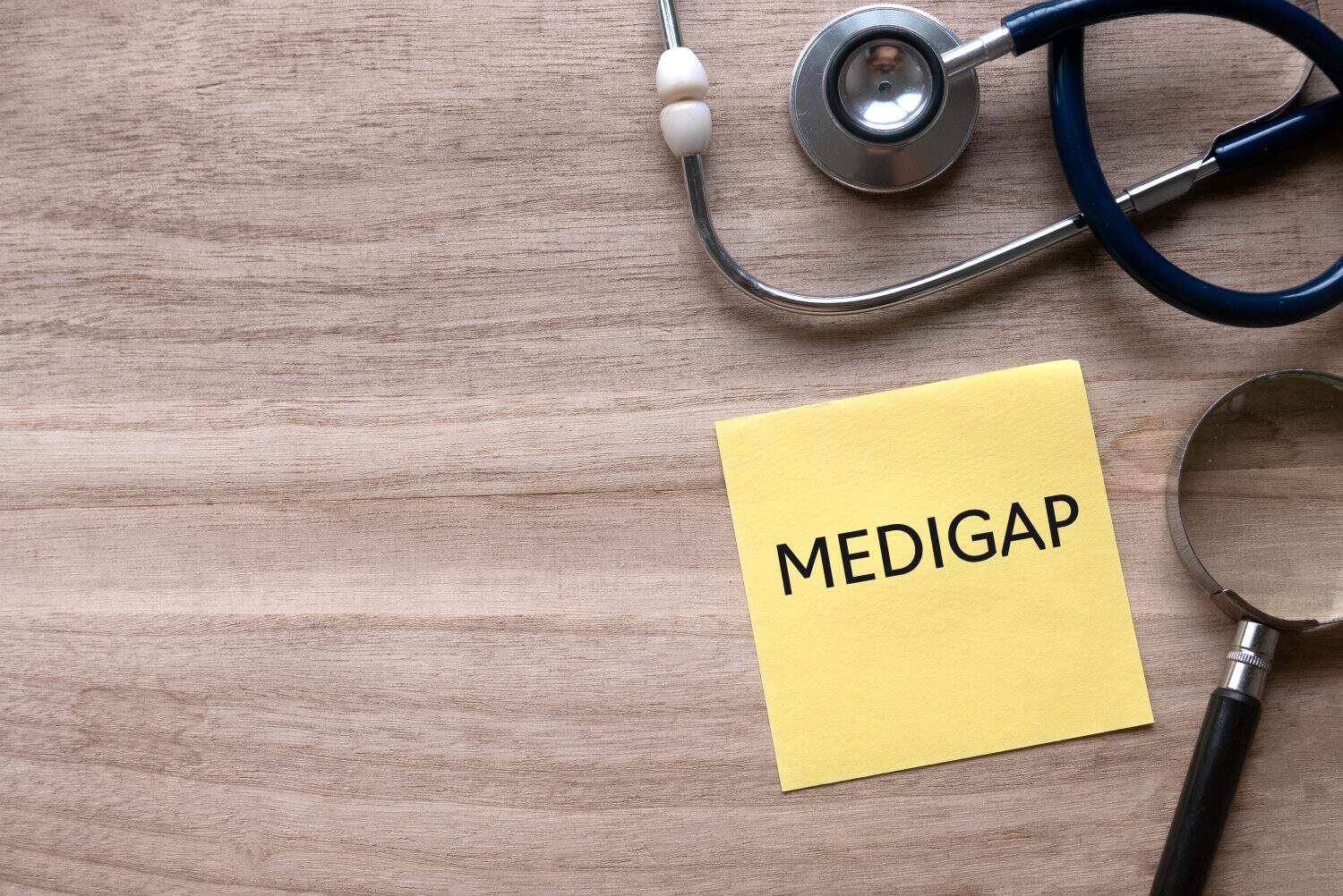 Top view of stethoscope, magnifying glass and yellow paper note written with Medigap on wooden background with copy space.