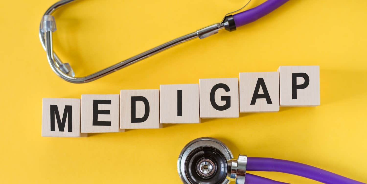 Word MEDIGAP building from wooden cubes on yellow desk with stethoscope.