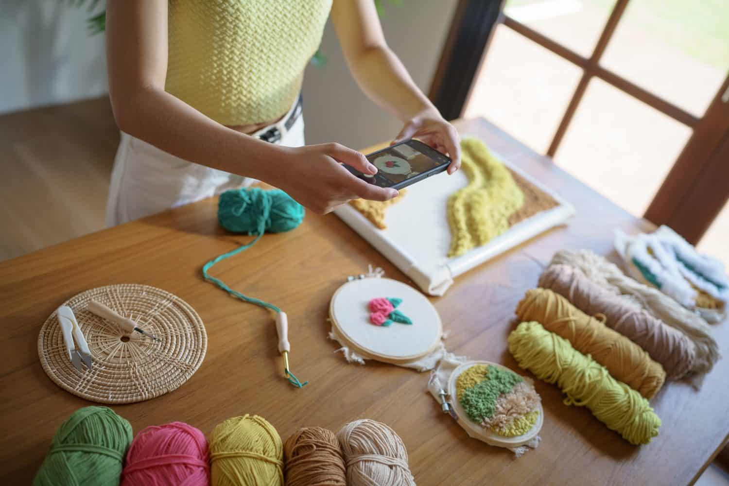 Asian Woman taking smartphone photo Punch needle. phone posting on social networks in studio workshop. designer workplace Handmade craft project DIY embroidery.