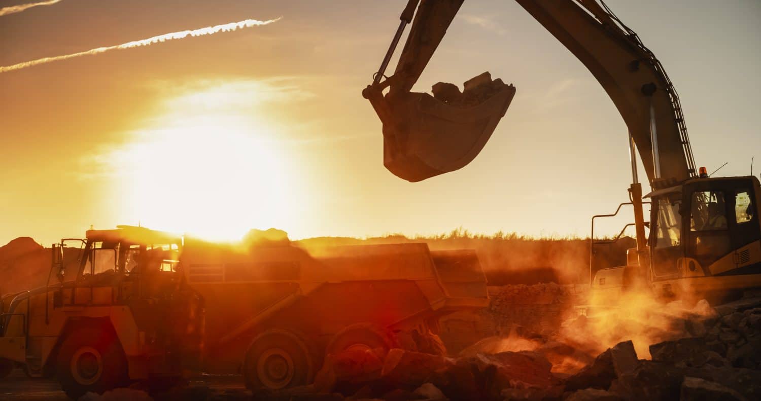 Construction Site On Sunny Evening: Industrial Excavator Loading Rocks Into A Truck. The Process Of Building New Apartment Complex. Workers Operating Heavy Machinery To Complete A Real Estate Project.