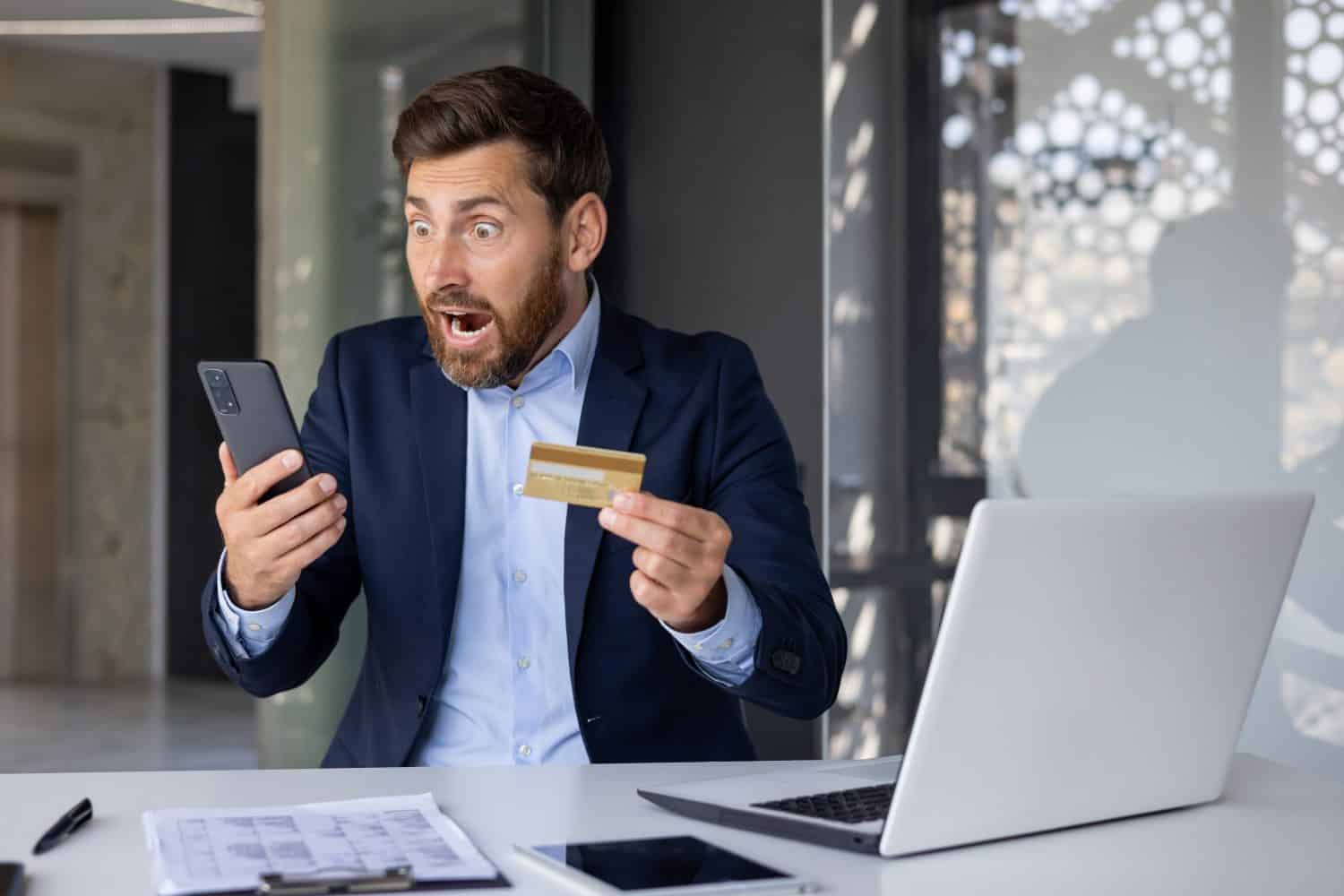 Shocked young man in business suit sitting at desk in office, holding credit card in hand and looking surprised at mobile phone screen.