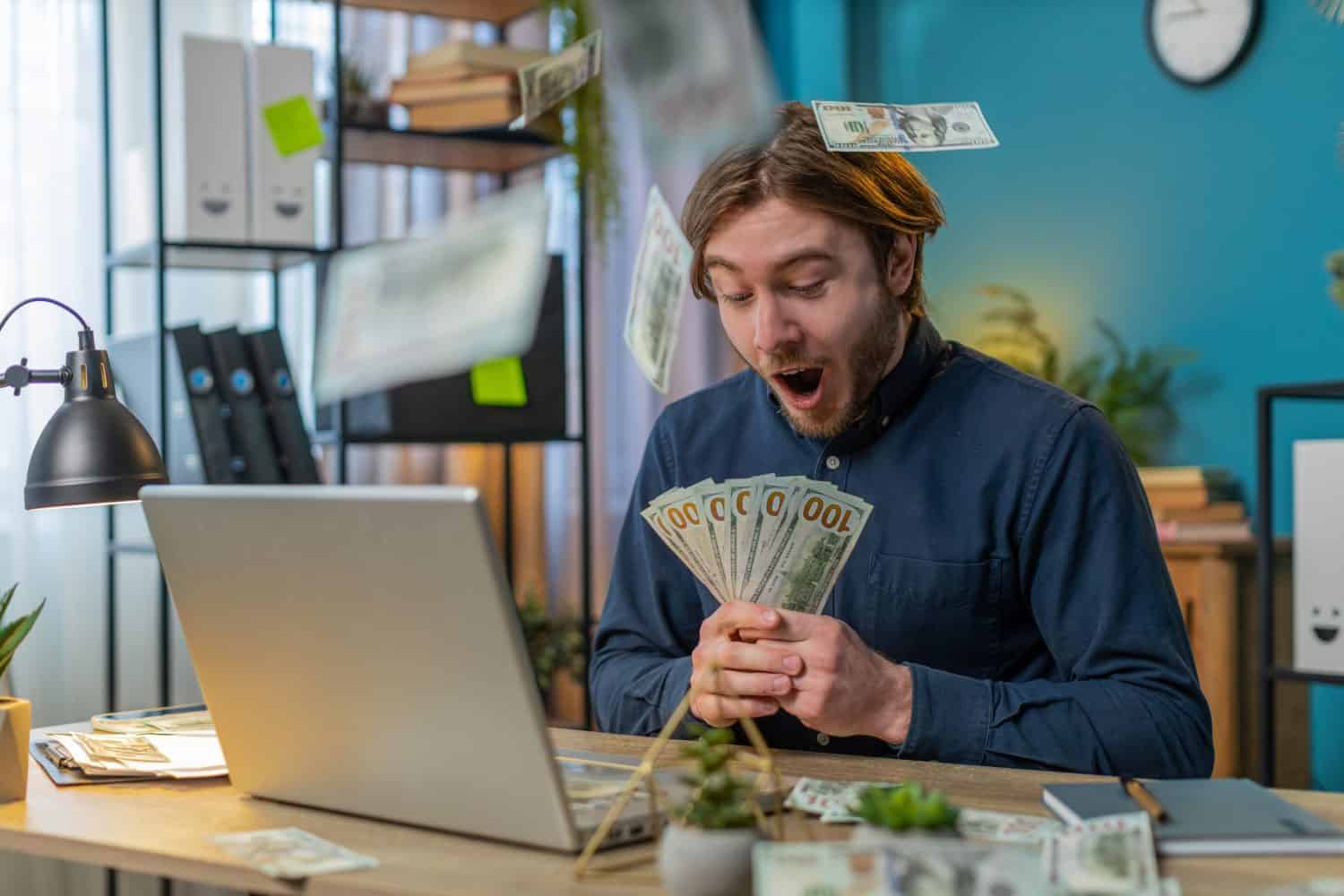 Rich Caucasian businessman earnings big profit success win lottery money cash rain falling shopping more tips. Happy young freelancer man guy manager with laptop clenching fists at home office desk