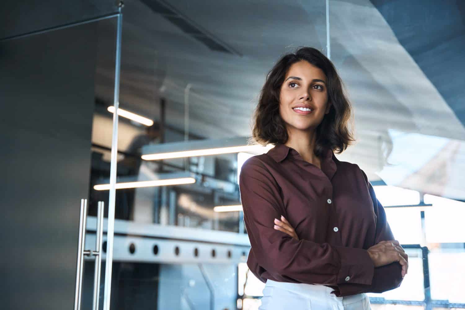 Latin or eastern middle age female ceo leader businesswoman standing in office. Portrait of beautiful successful hispanic young business woman with crossed arms looking confident aside. Copy space