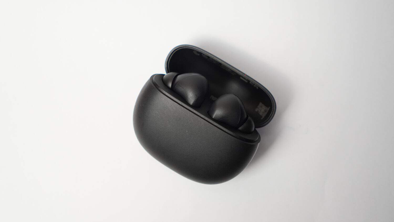 A sleek black wireless earbud case with two earbuds inside is displayed on a white background. The case is slightly open, revealing the earbuds securely nestled in their slots. The minimalist design e