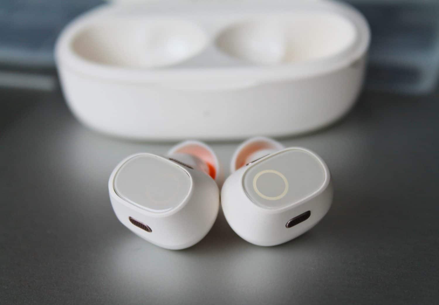 A pair of white wireless earbuds with indicator ring lights on. And a blurry charging case in the background. Closeup.