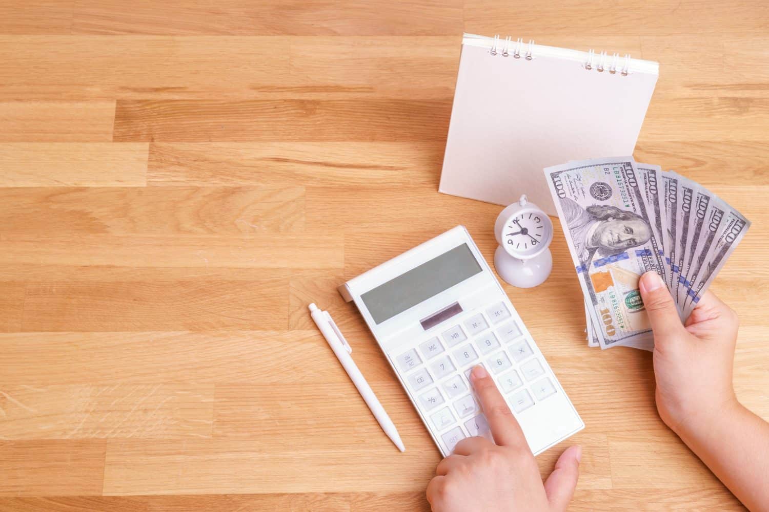 Hands using white modern design calculator and holding dollar cash with pen, clock and calendar on light wooden table background for saving money and salary planning concept captured in top view