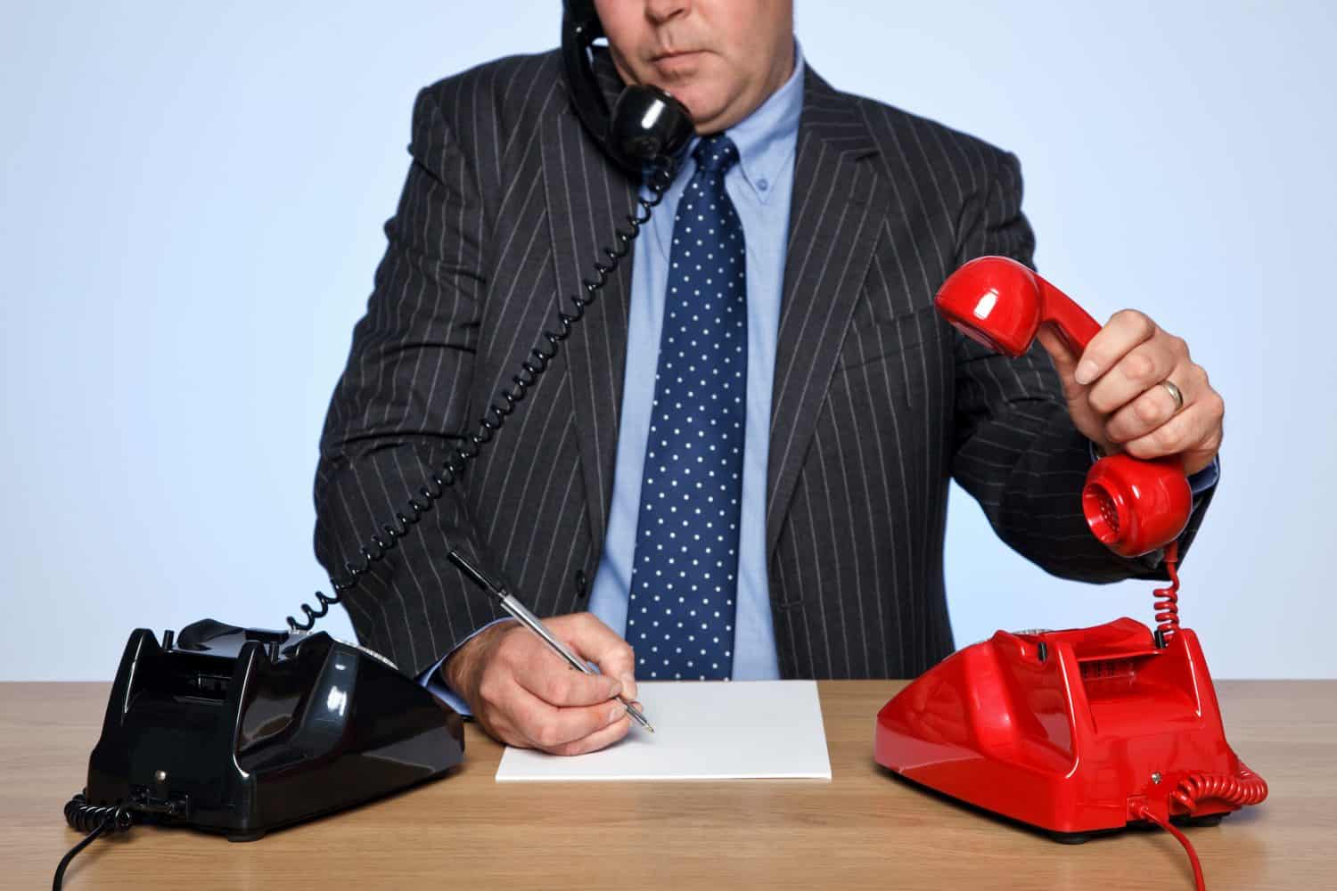 Photo of a businessman sat at a desk with two traditional telephones, one red and one black. He is listening to one call whilst picking up the other phone.