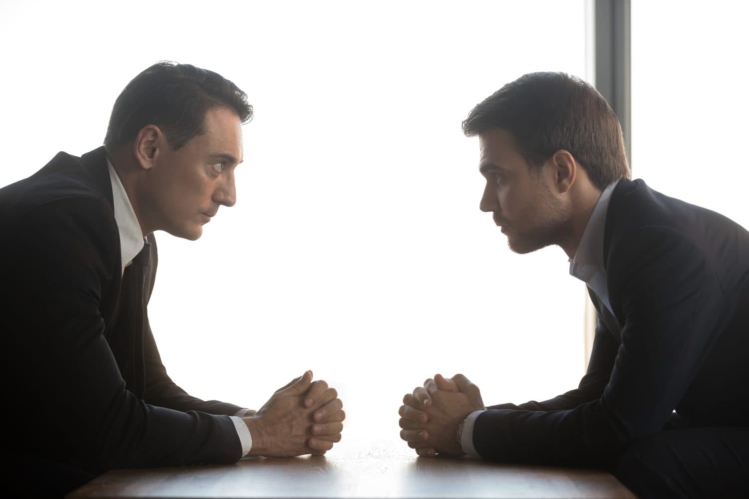 Two confident businessmen with clasped hands look at each other sitting opposite as rivalry confrontation concept, business opponents competitors politicians debate, difficult negotiations, side view