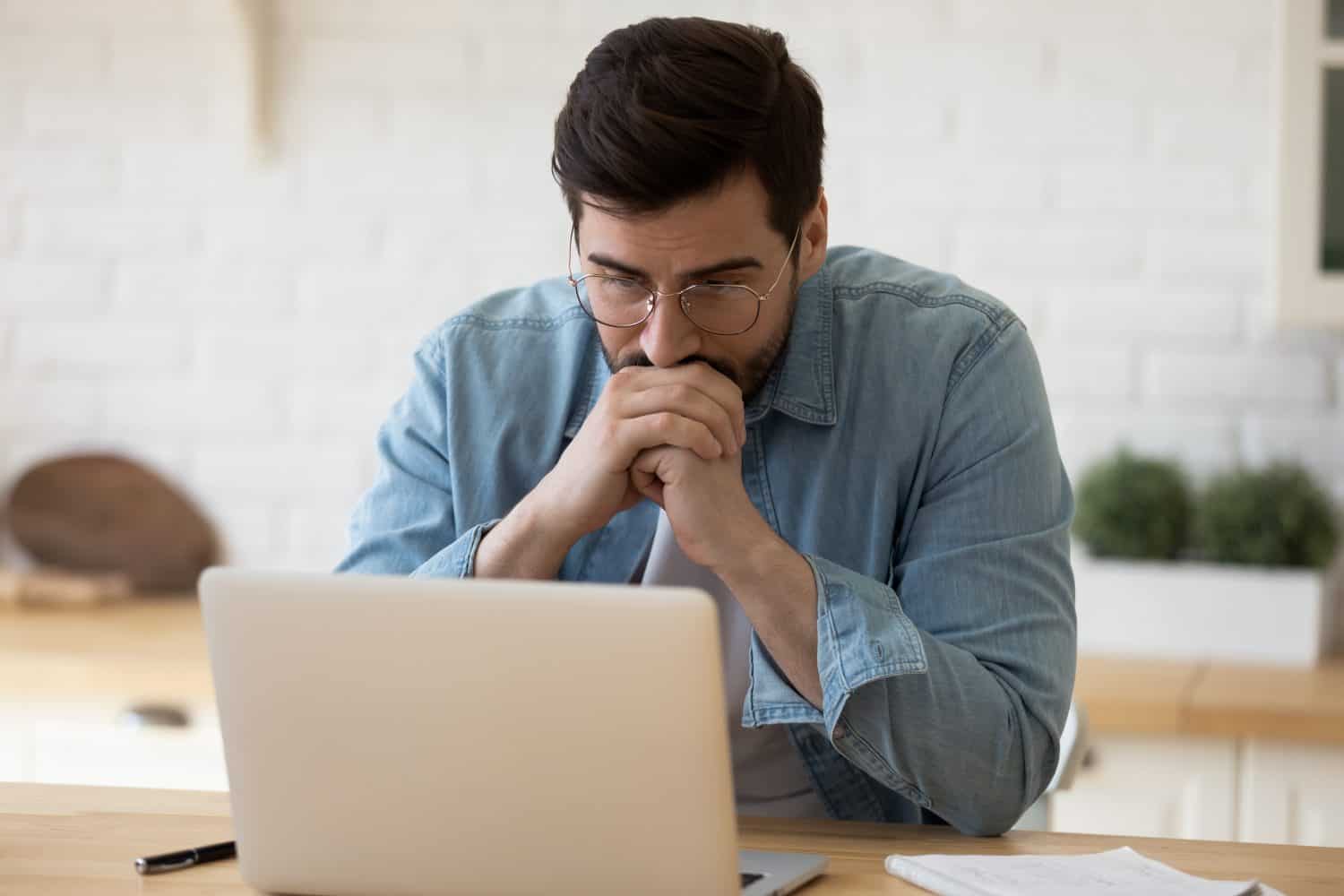 Head shot thoughtful businessman in eyewear looking at computer screen, sitting at table at home. Pensive confused young man thinking of problem solution stuck with task, working with laptop remotely.