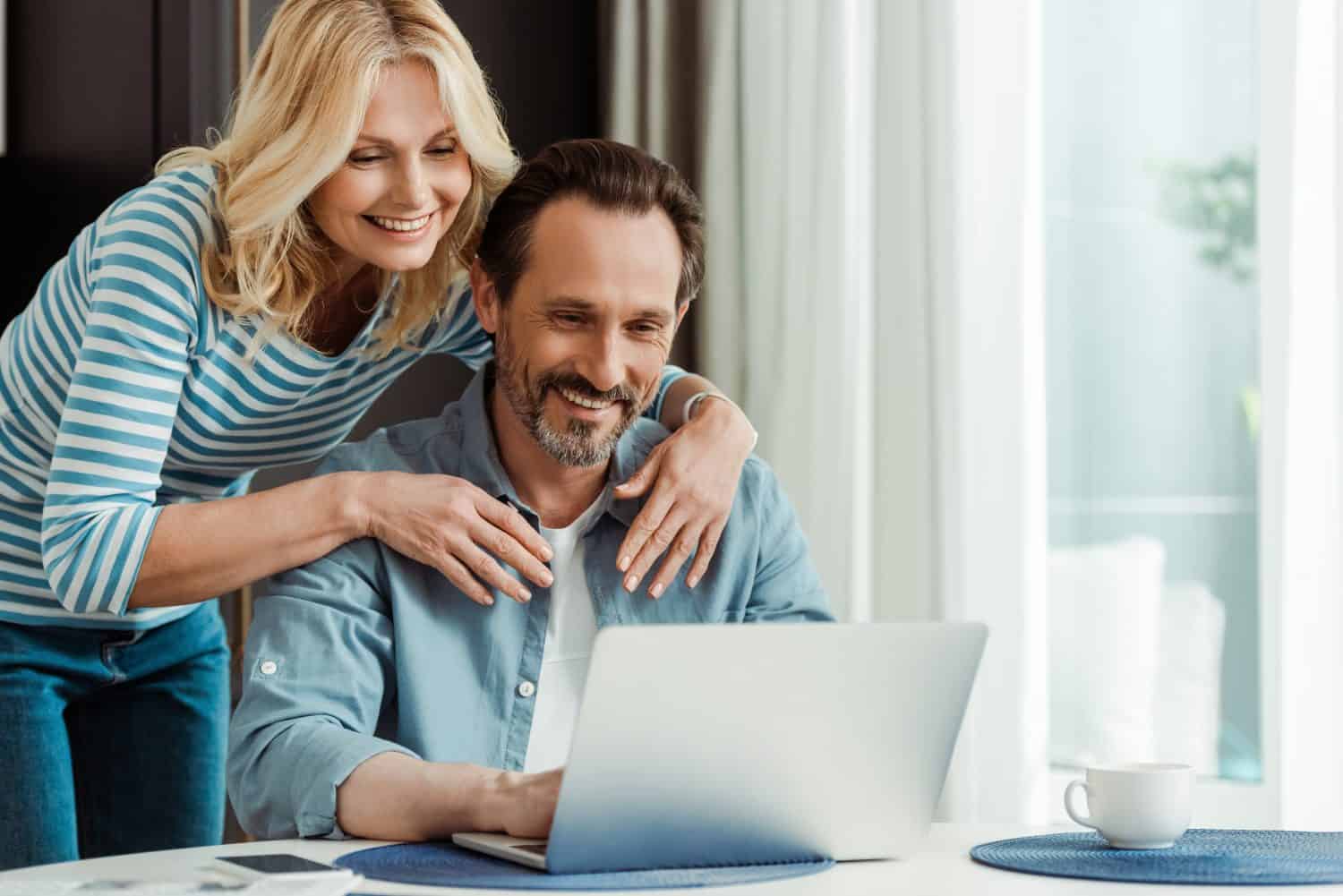 Selective focus of smiling woman embracing husband using laptop in kitchen