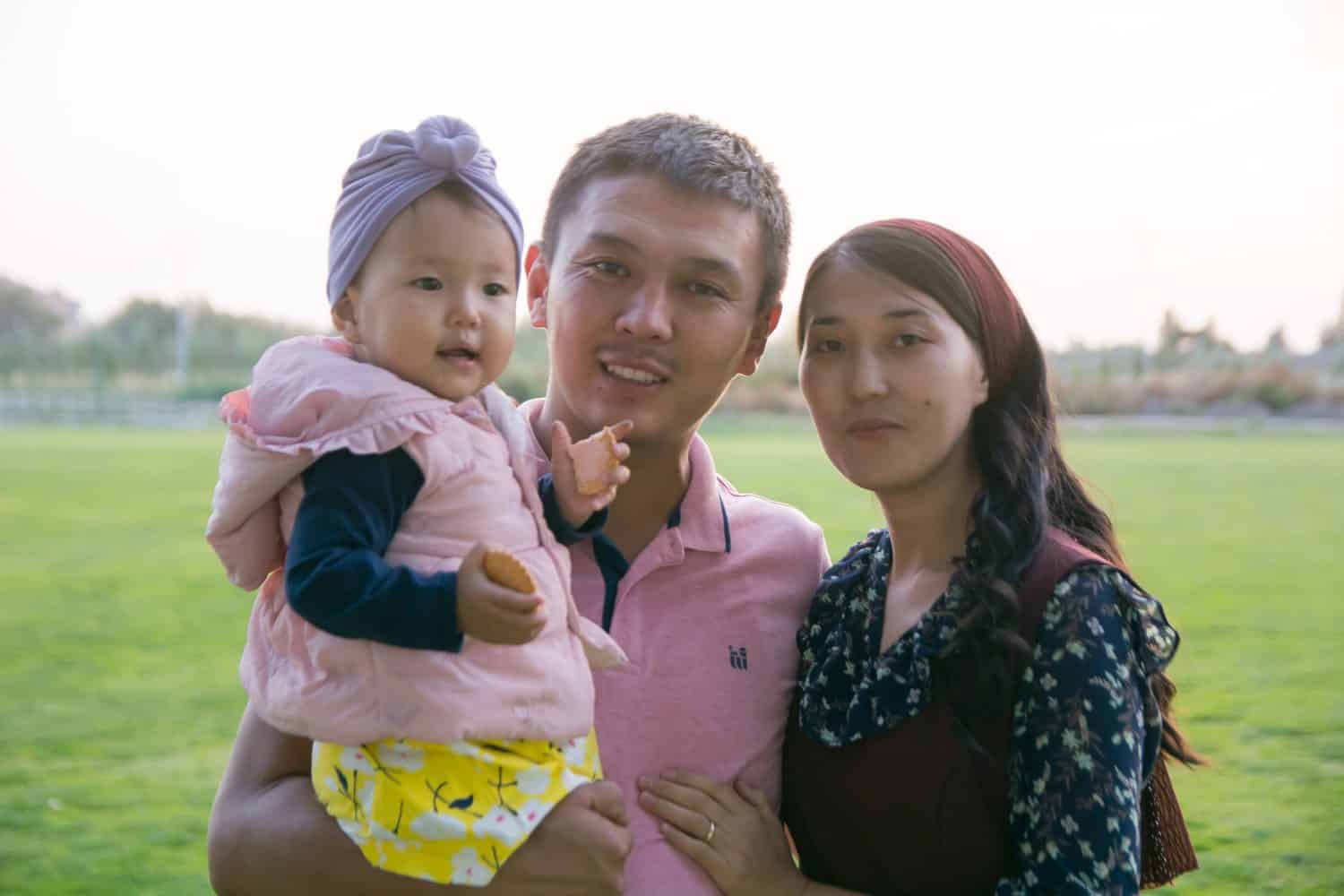 Parents hold their daughter in their arms, walking along the green lawn in the park.