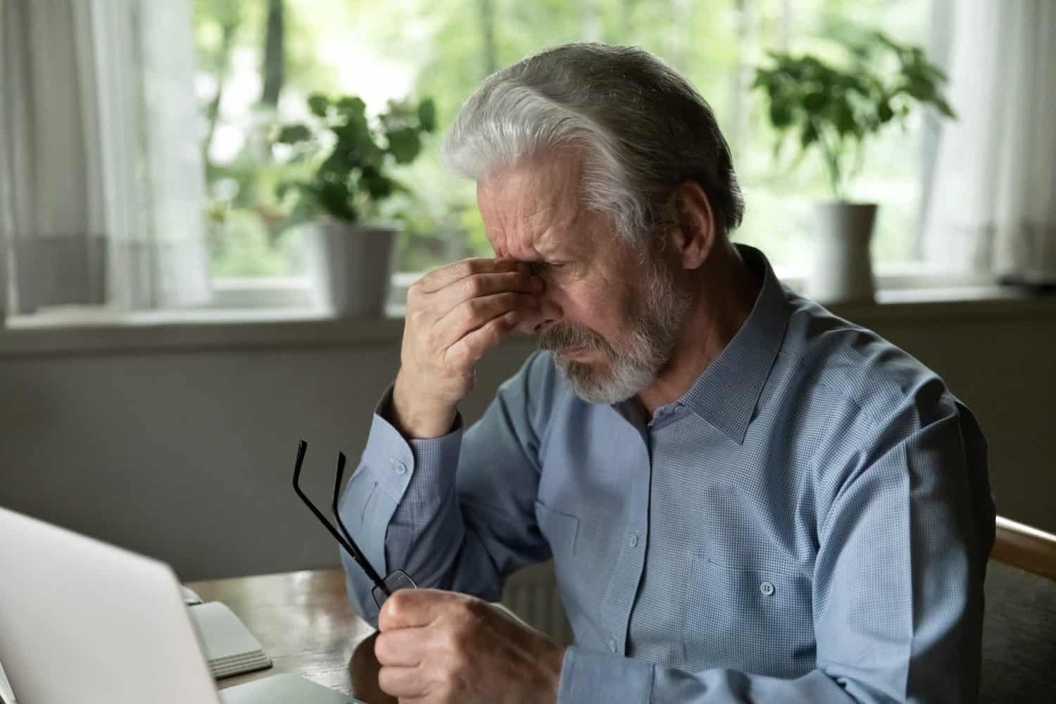 Close up stressed mature man massaging nose bridge, holding glasses, sitting at desk with laptop, elderly grey haired male suffering from eye strain, dry eyes syndrome after long laptop use