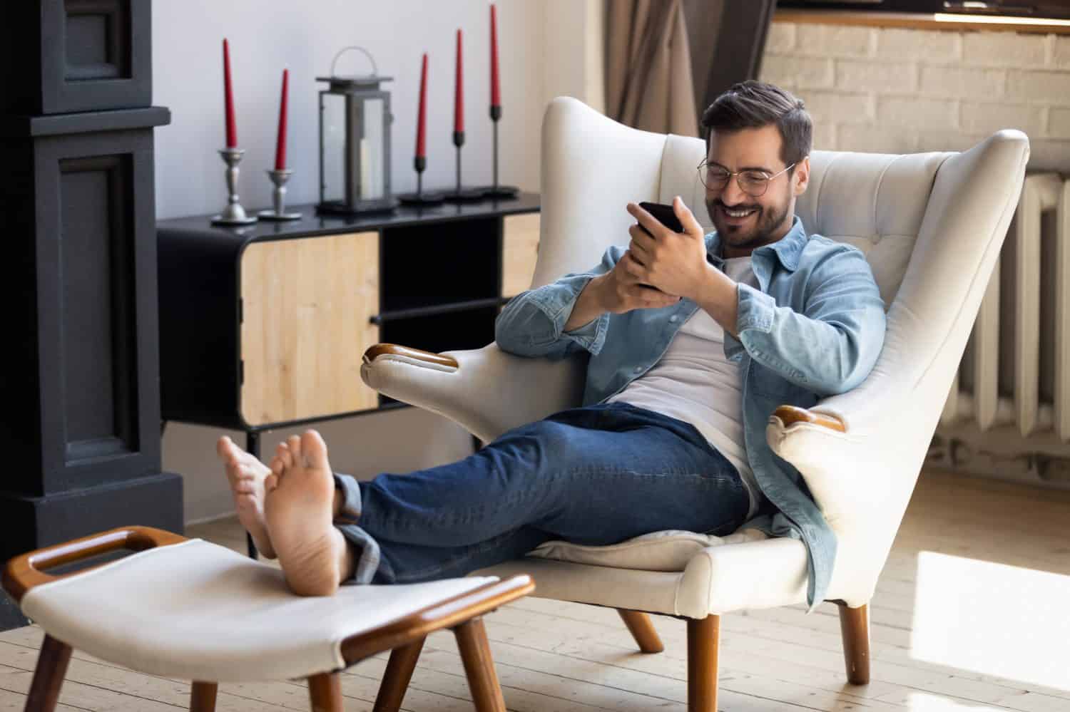 Handsome 30s single man relaxing on comfy armchair put feet on footstool holding in hands smartphone spend weekend alone in cozy cottage house. No stress, home owner, modern tech usage, fun concept