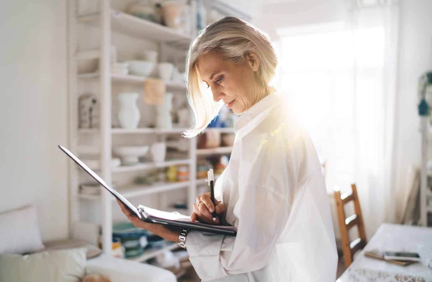 Focused mature european female entrepreneur writing something in clipboard. Small business and entrepreneurship. Modern successful woman. Home art studio with pottery on shelves at sunny day