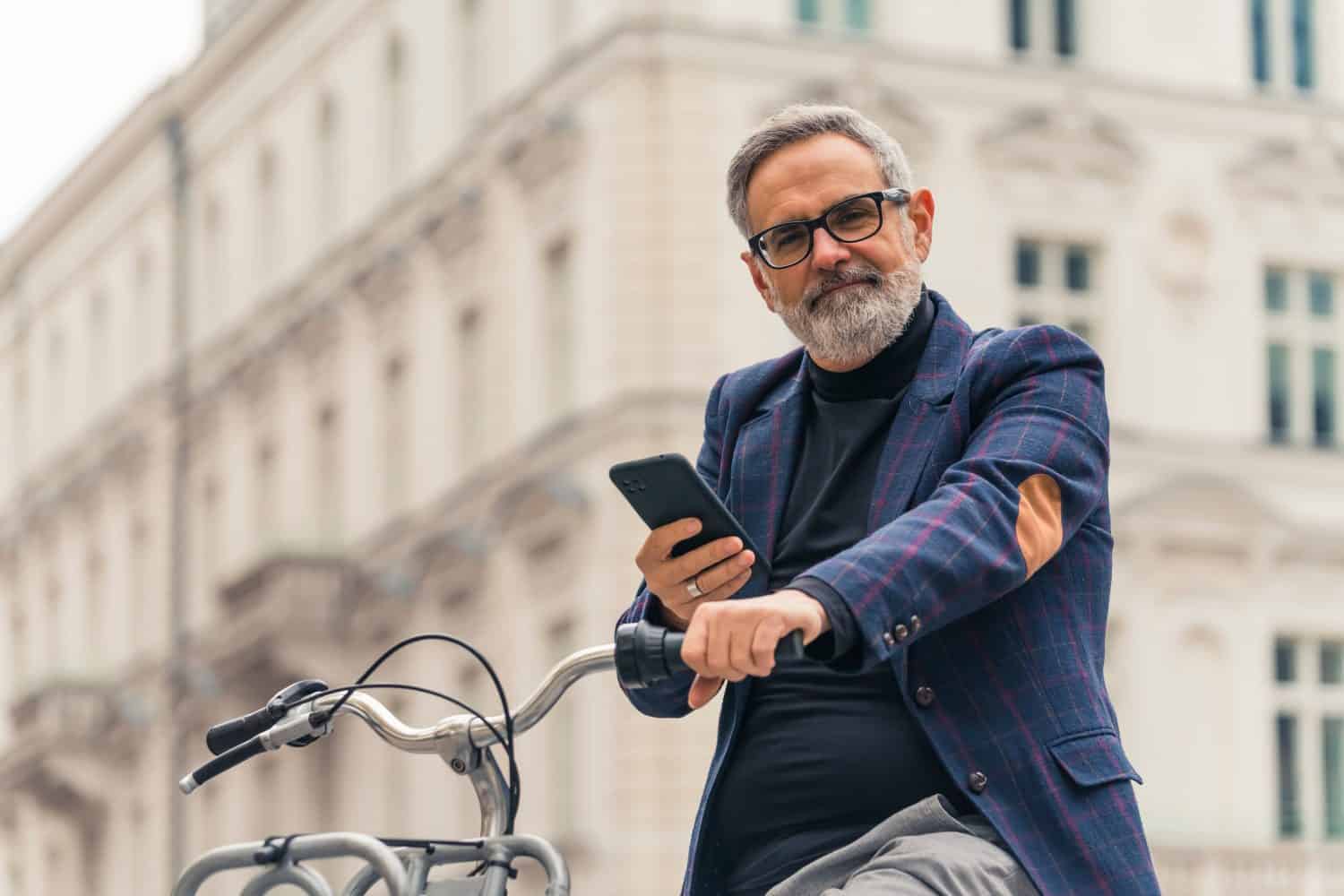 60-year-old caucasian gray-haired bearded businessman spending his break outdoors, looking at camera while holding bike wheel and big smartphone in black case. Blurred white elegant building in the