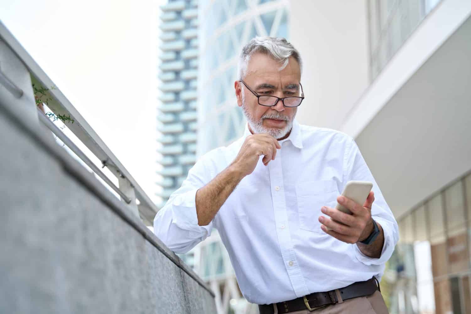 Older professional business man, serious senior 60 years old businessman holding smartphone using mobile cell phone technology and thinking standing outdoor in big city office district.