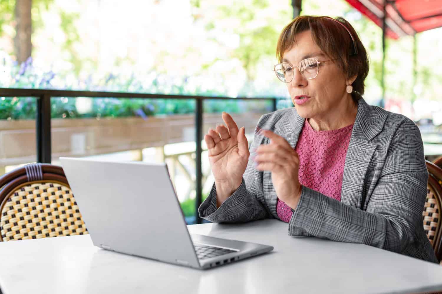 Photo of a serious 60-years-old woman is online working at summer cafe. She sitting in front of laptop monitor and talking during online video call. Modern technology in every day life concept.