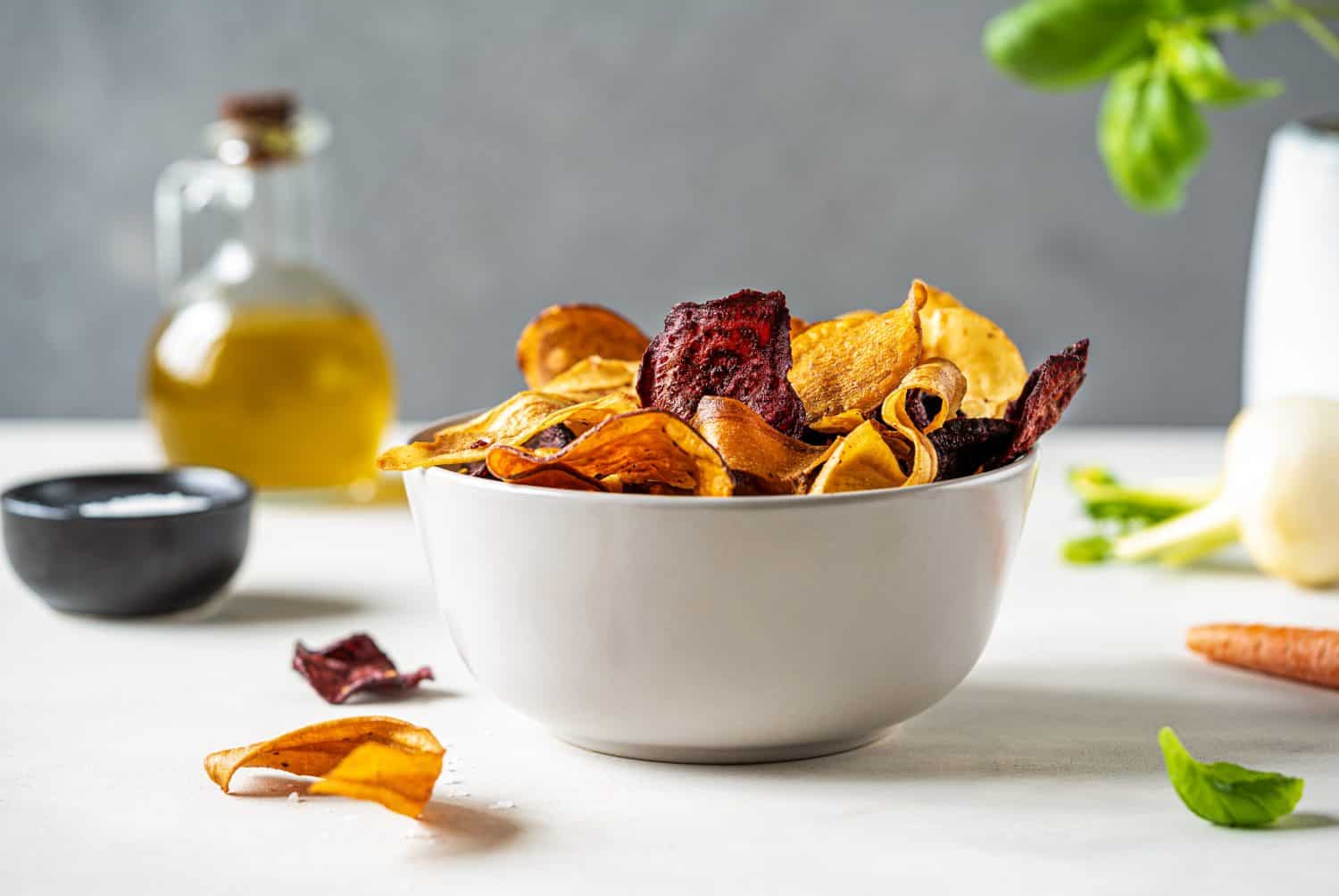 Vegetable chips in a bowl on light surface with grey background, salt, basil, vegetables, oil