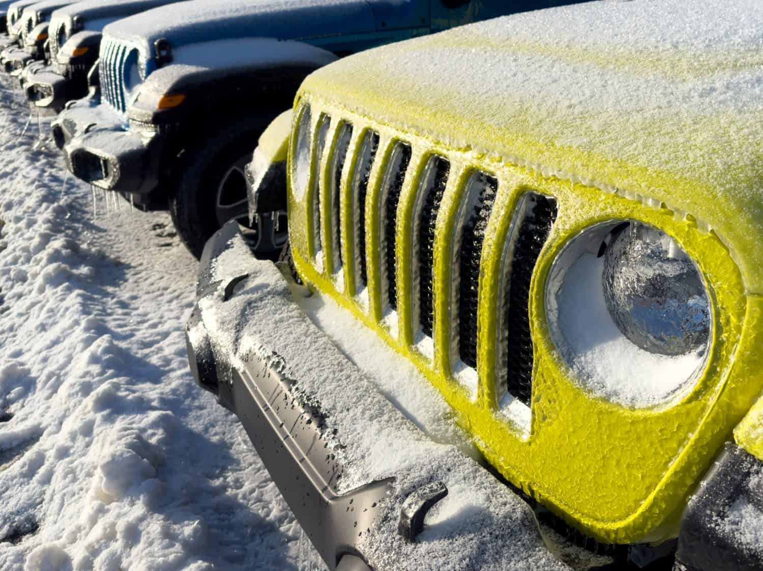 A row of new 2024 Jeep 4x4 sporty Wrangler SUVs and Gladiator trucks in a line. Some are bright yellow, blue, black, and green. The recreation vehicles are covered in thick winter ice and frost snow.