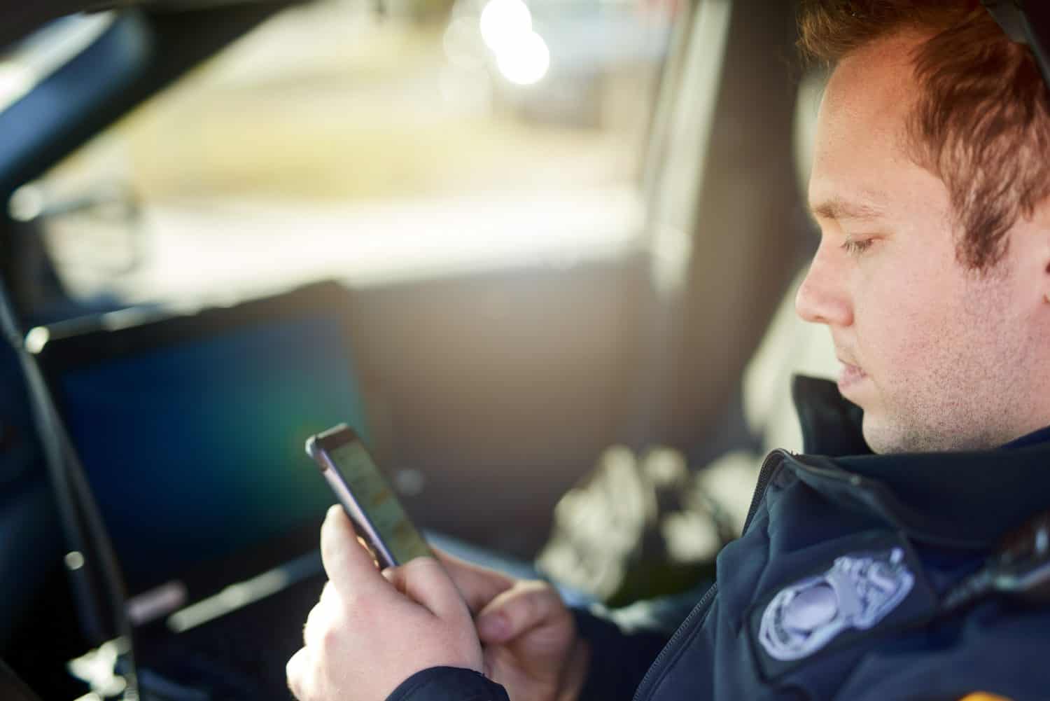 Police, officer and man texting with phone in patrol car for security contact, law enforcement update and mobile notification. Policeman typing message on cellphone for crime news, connection and app