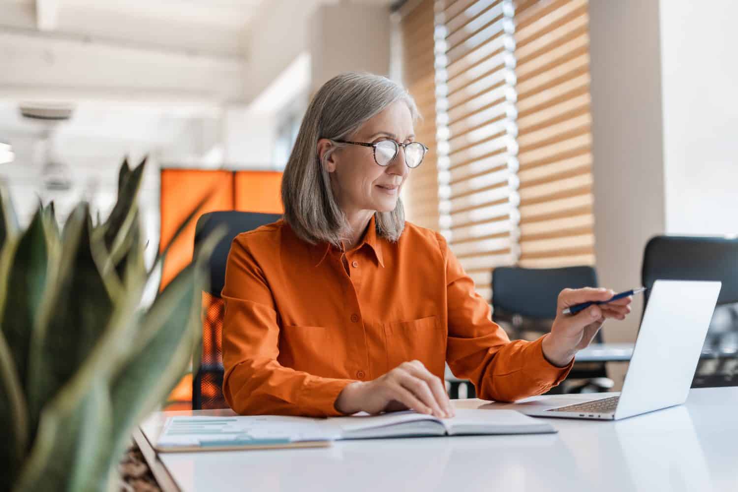 Portrait of beautiful confident senior woman, manager using laptop computer working online sitting in modern office. Business woman wearing stylish eyeglasses checking email. Technology concept