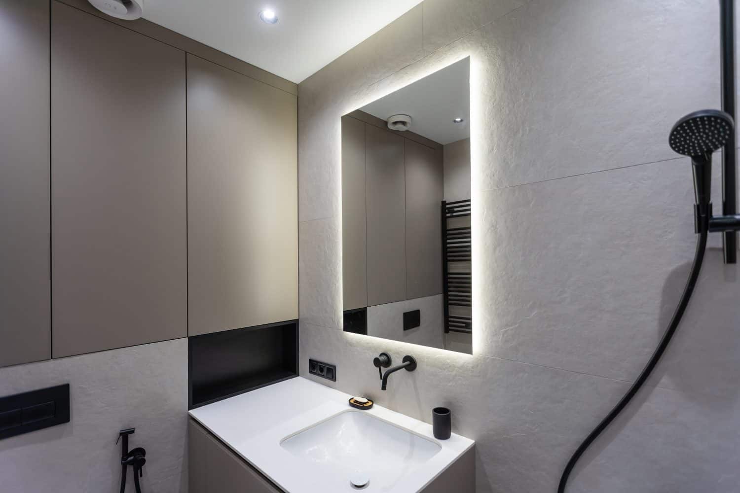 Bathroom in a modern style with gray and white tiles. There is a large mirror with luminous lamps, tabletop with wooden drawers and sink, bath with shower and glass partition, towel rack and a hanger.