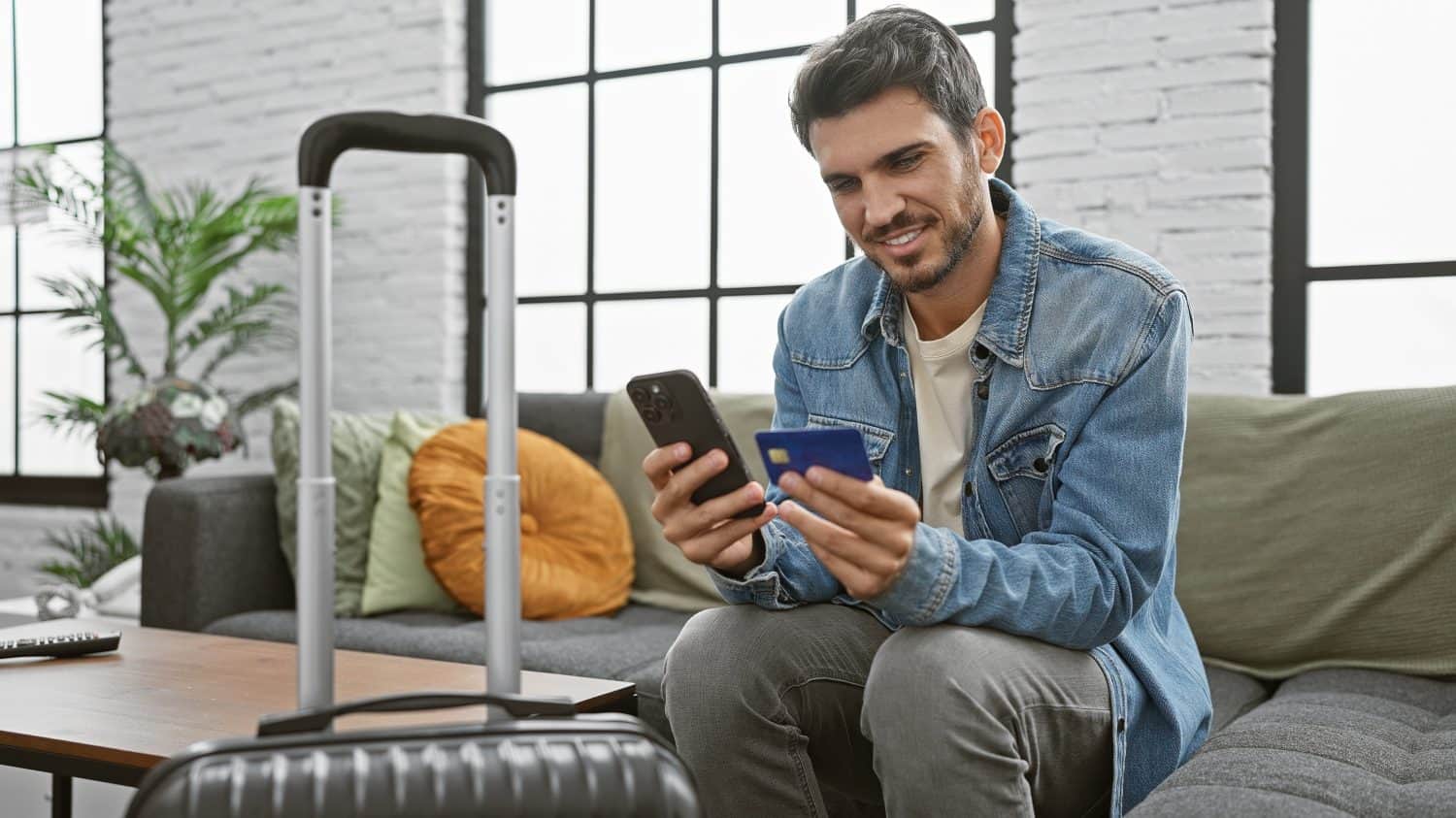 Handsome hispanic man with beard using smartphone and holding credit card in modern apartment living room with suitcase.