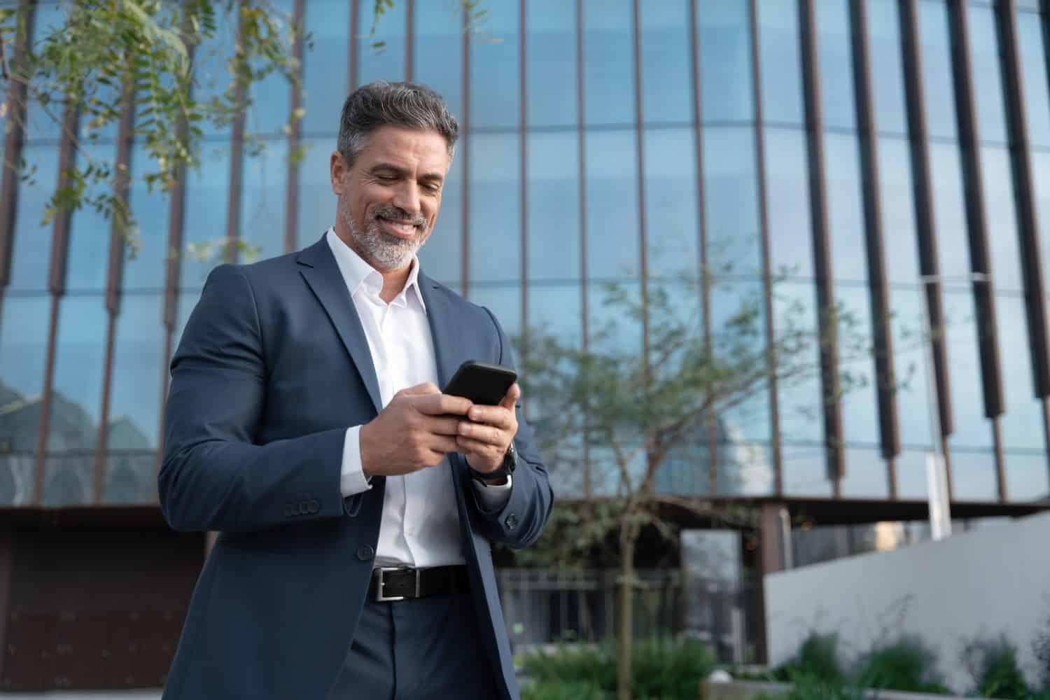 Smiling mature hispanic senior business man using smartphone cellphone at office building. Latin middle age male entrepreneur businessman holding mobile cell phone for trading outdoors. Copy space