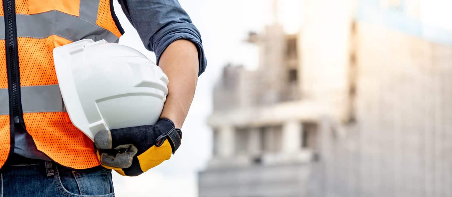 Safety workwear concept. Male hand holding white safety helmet or hard hat. Construction worker man with reflective orange vest and protective gloves standing at unfinished building with tower crane