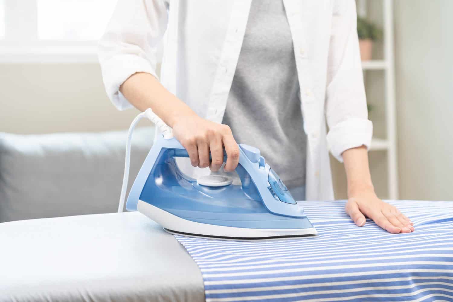 Close up young woman hand use electric steam, hot iron press pile stripe shirt clothes on ironing board, housework after hygiene laundry at home. Housekeeping lifestyle, household of chores concept.