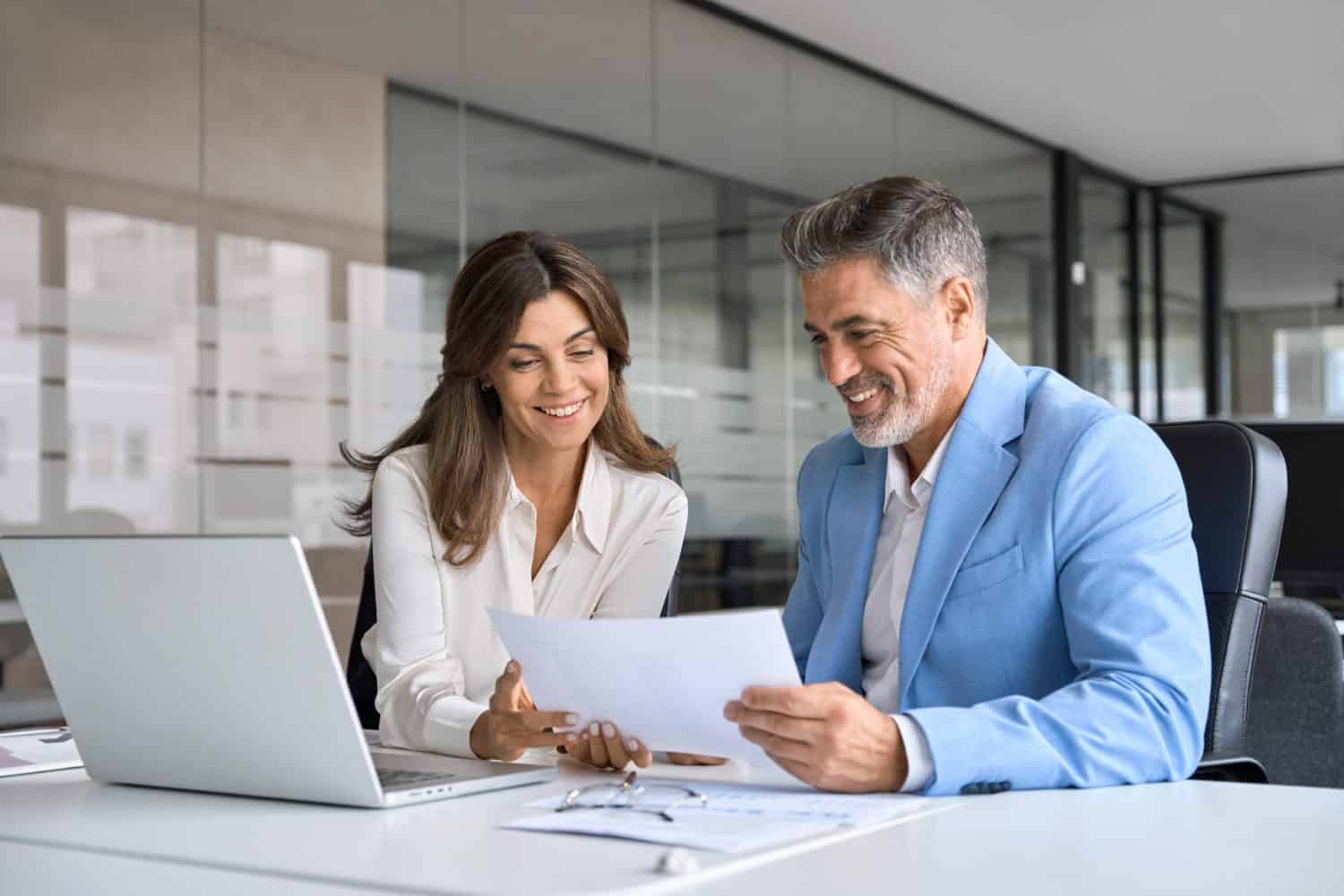 Two happy busy middle aged professionals man and woman business leaders partners checking document reading financial report talking working together on laptop computer in office at corporate meeting.