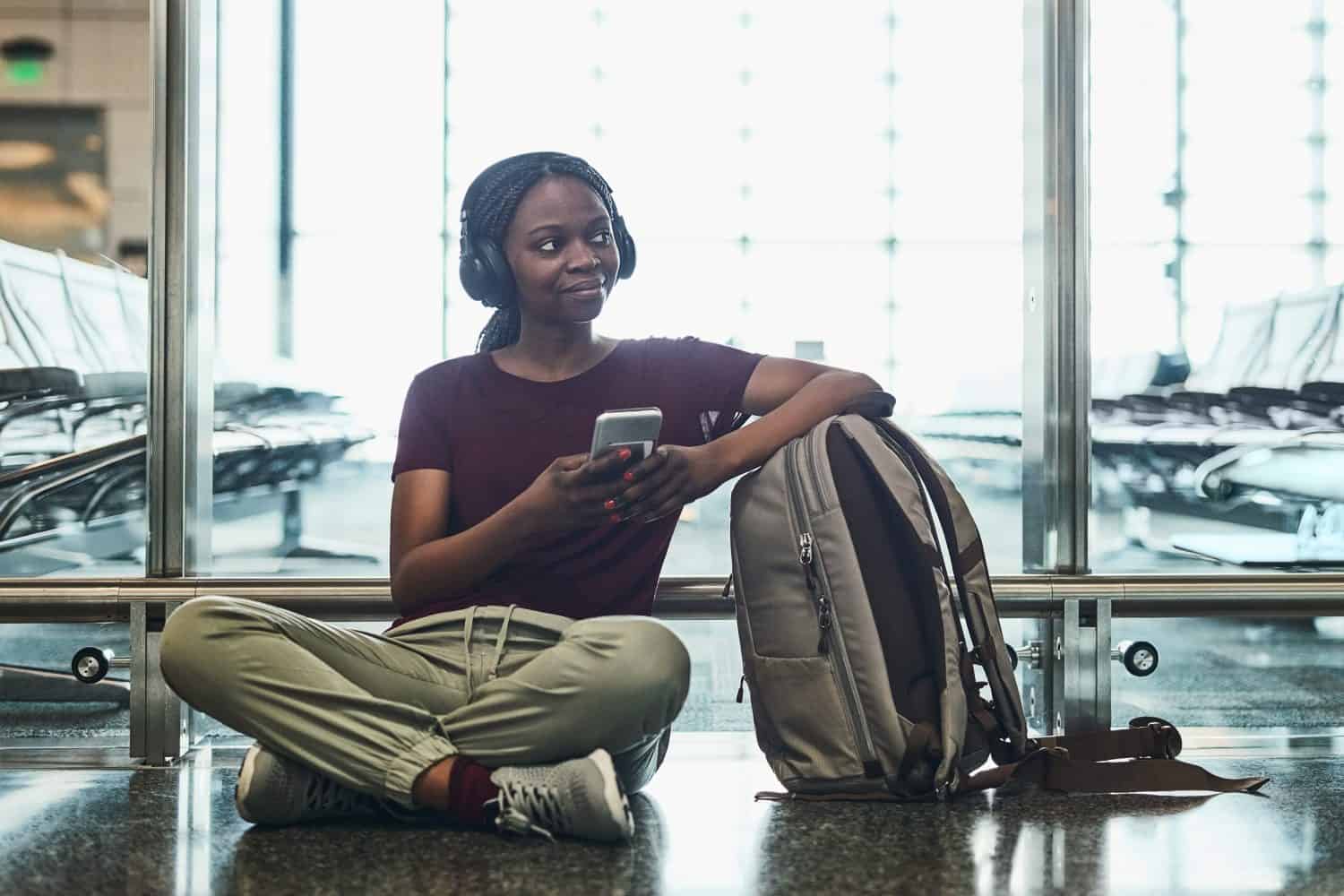 Music, headphones and black woman with phone at airport for audio, streaming and immigration. International, travel and person with tech on floor for network, connection and online flight information