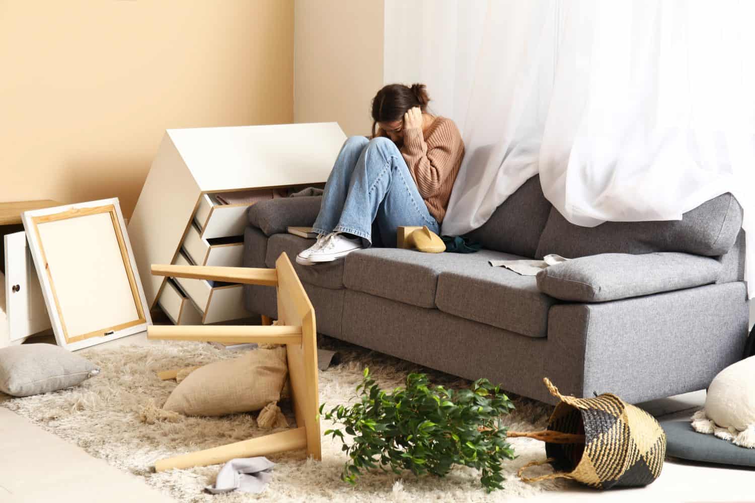 Young woman sitting on sofa in messed living room after earthquake