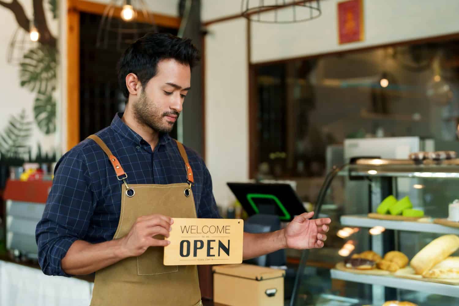 Concentrated barista holding &#039;Open&#039; sign beside counter, pastry showcase slightly visible. Coffee shop attendant in trendy workwear with welcoming gesture, baked goods in soft focus