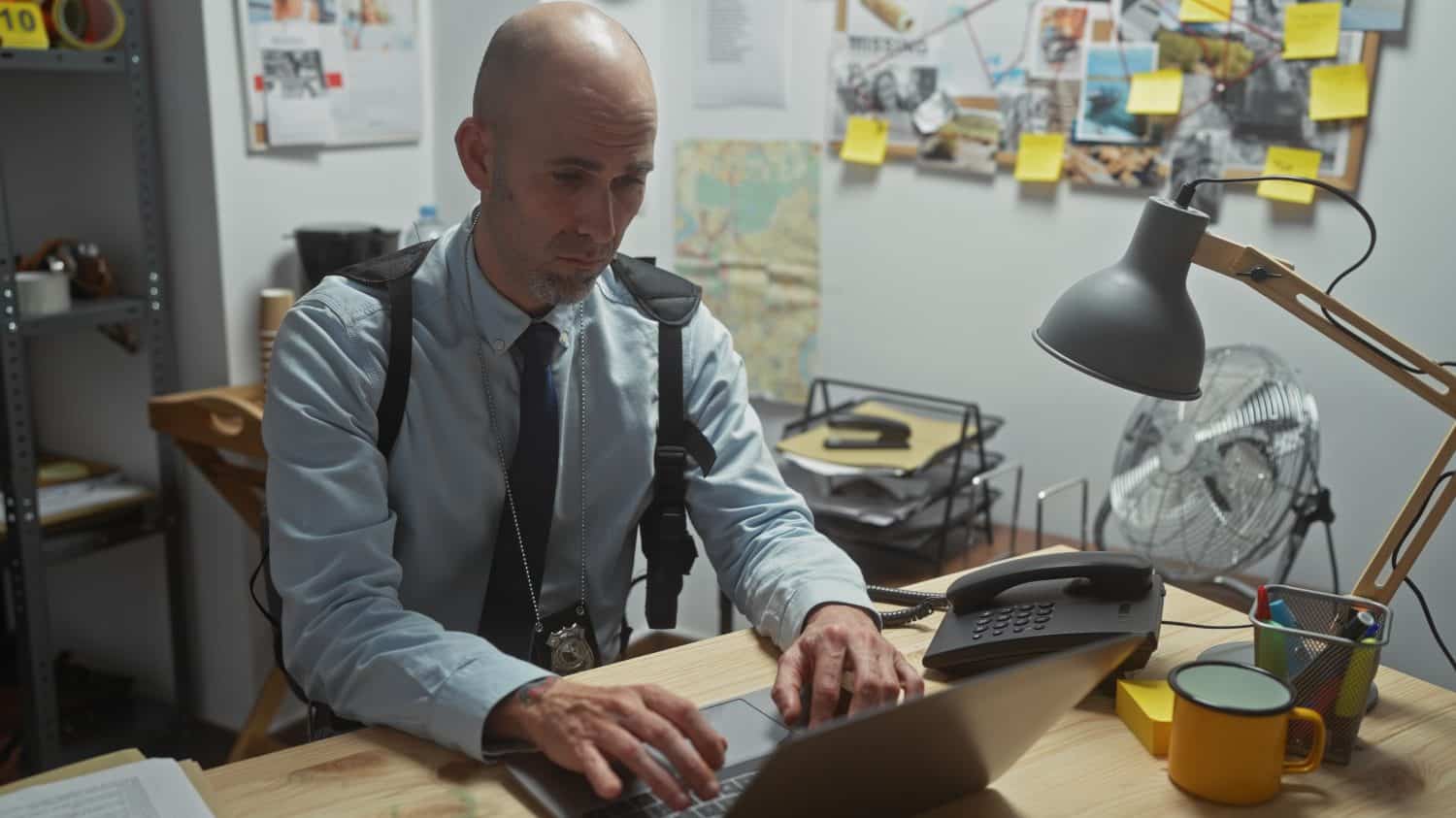 A bald man with a beard working on a laptop in a cluttered detective office surrounded by evidence boards.