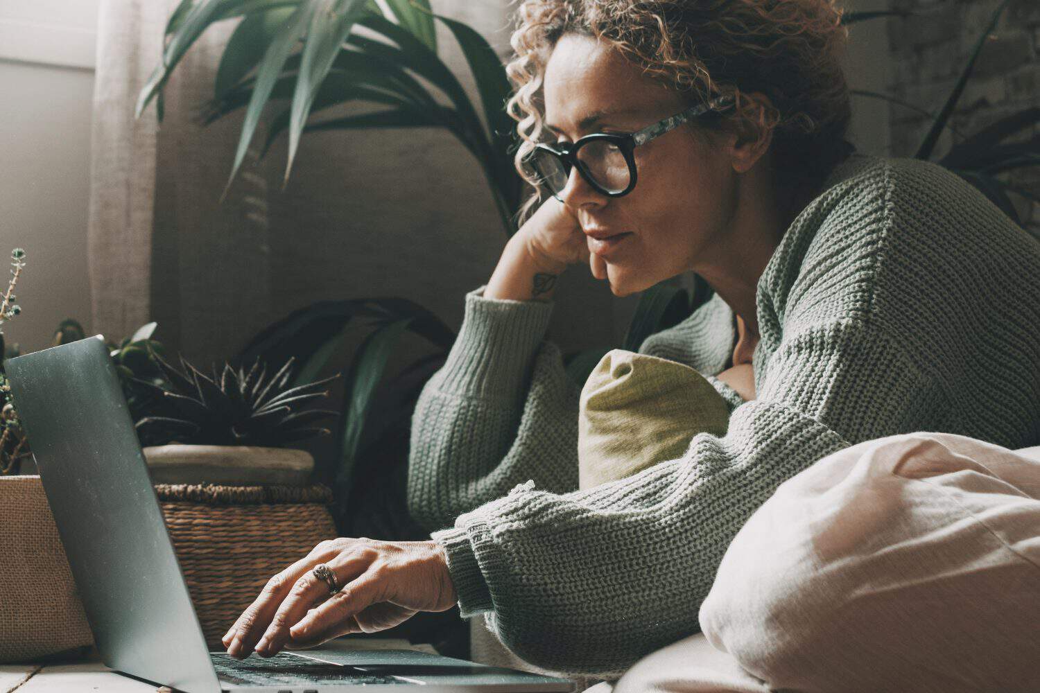 Blonde woman with curly hair using laptop sitting on sofa at home. 45 year old serious middle aged female looking at computer while surfing internet, shopping on website and relaxing on sofa.