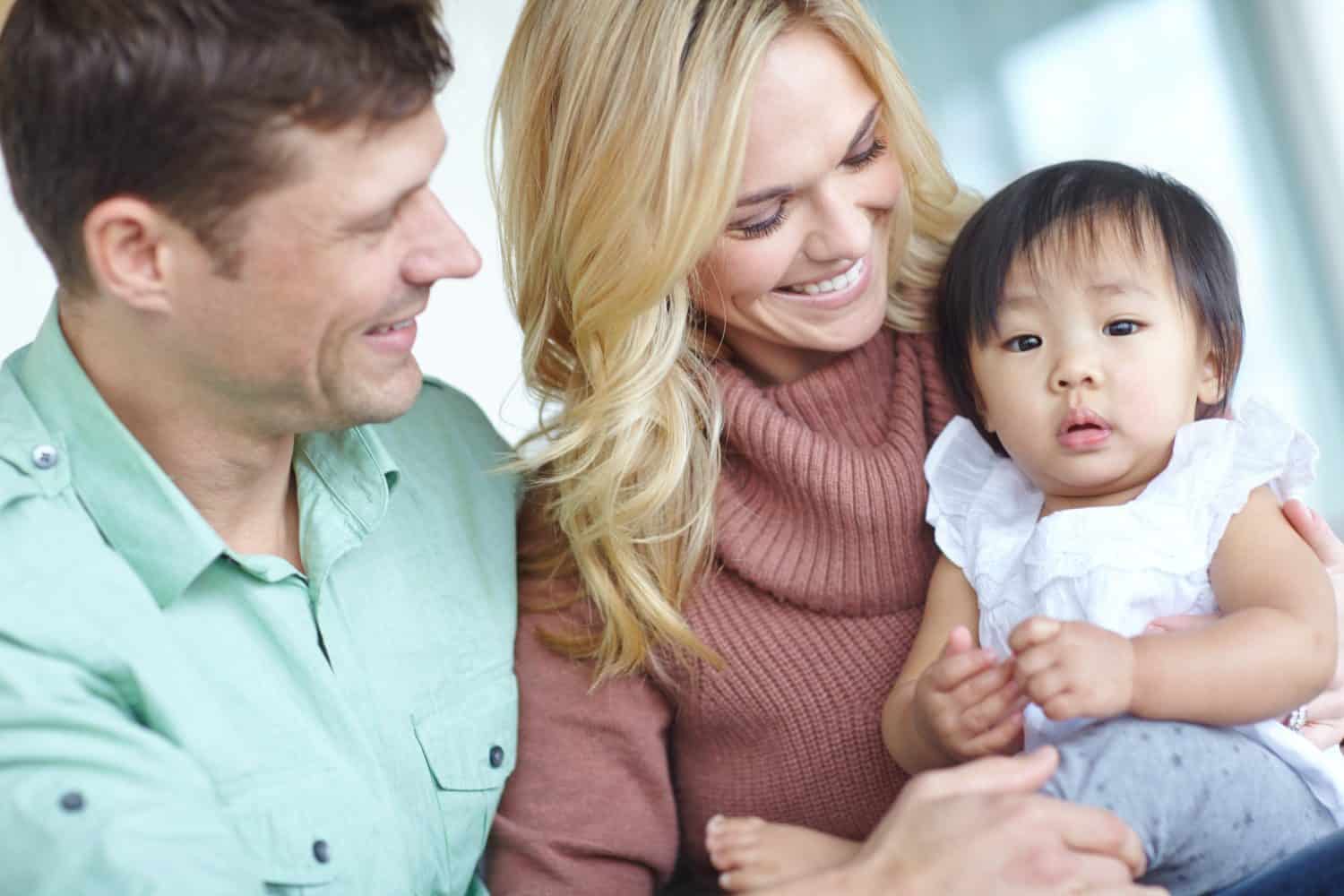 Couple, adoption and asian child for family in home, love or girl for foster care. Together, bonding or support with married man and female person, diversity and baby for trust and hug in living room