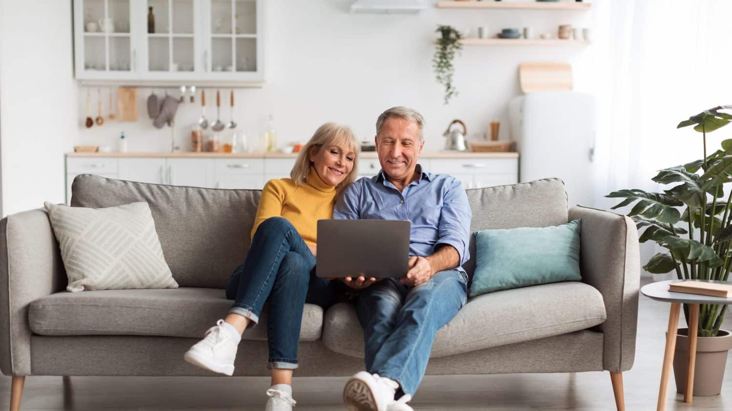 Senior Spouses Using Laptop Watching Movie Together Sitting On Couch At Home. Older Couple Browsing Internet On Computer Reading Online News On Weekend. Technology And Gadgets