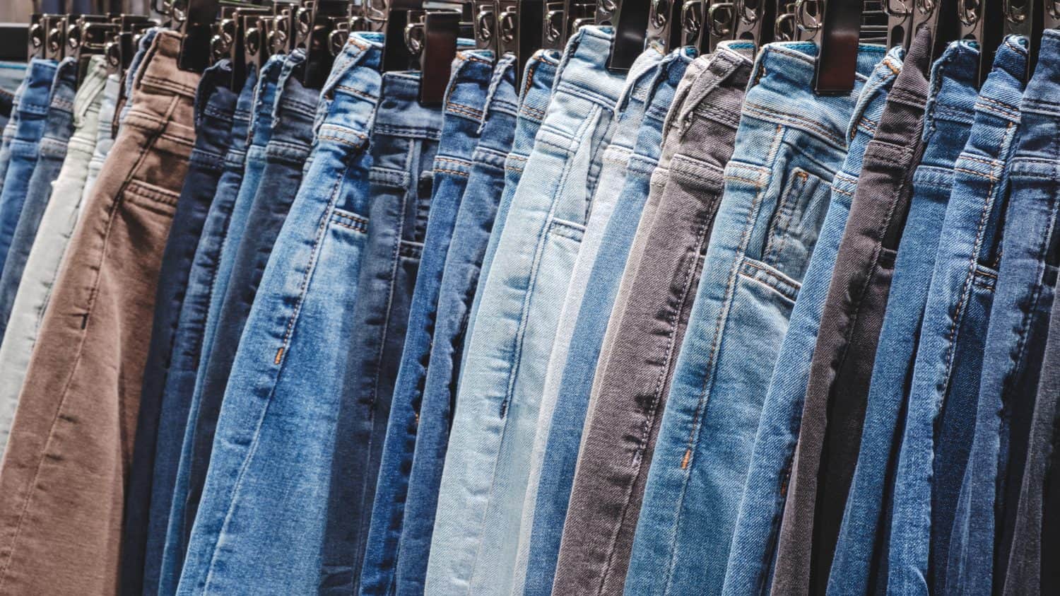 A row of jeans hanging on a rack. The jeans are of different colors and styles. Concept of variety and choice, as the person hanging the jeans can pick from a wide range of options