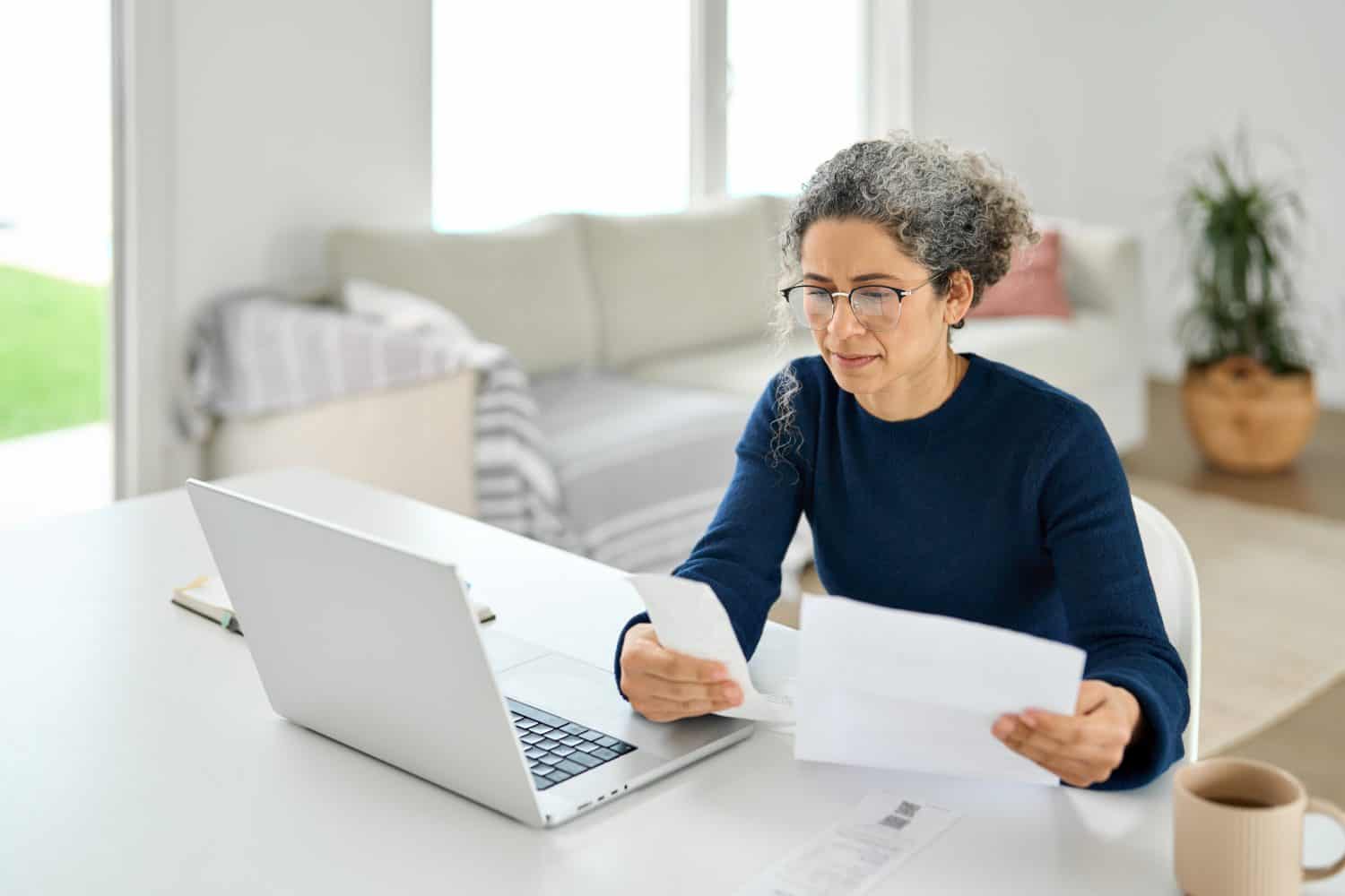 Mature lady paying bills online checking medical invoices receipts bills making baking payments or calculating tax refund using laptop. Middle aged older woman worrying about financial problem at home