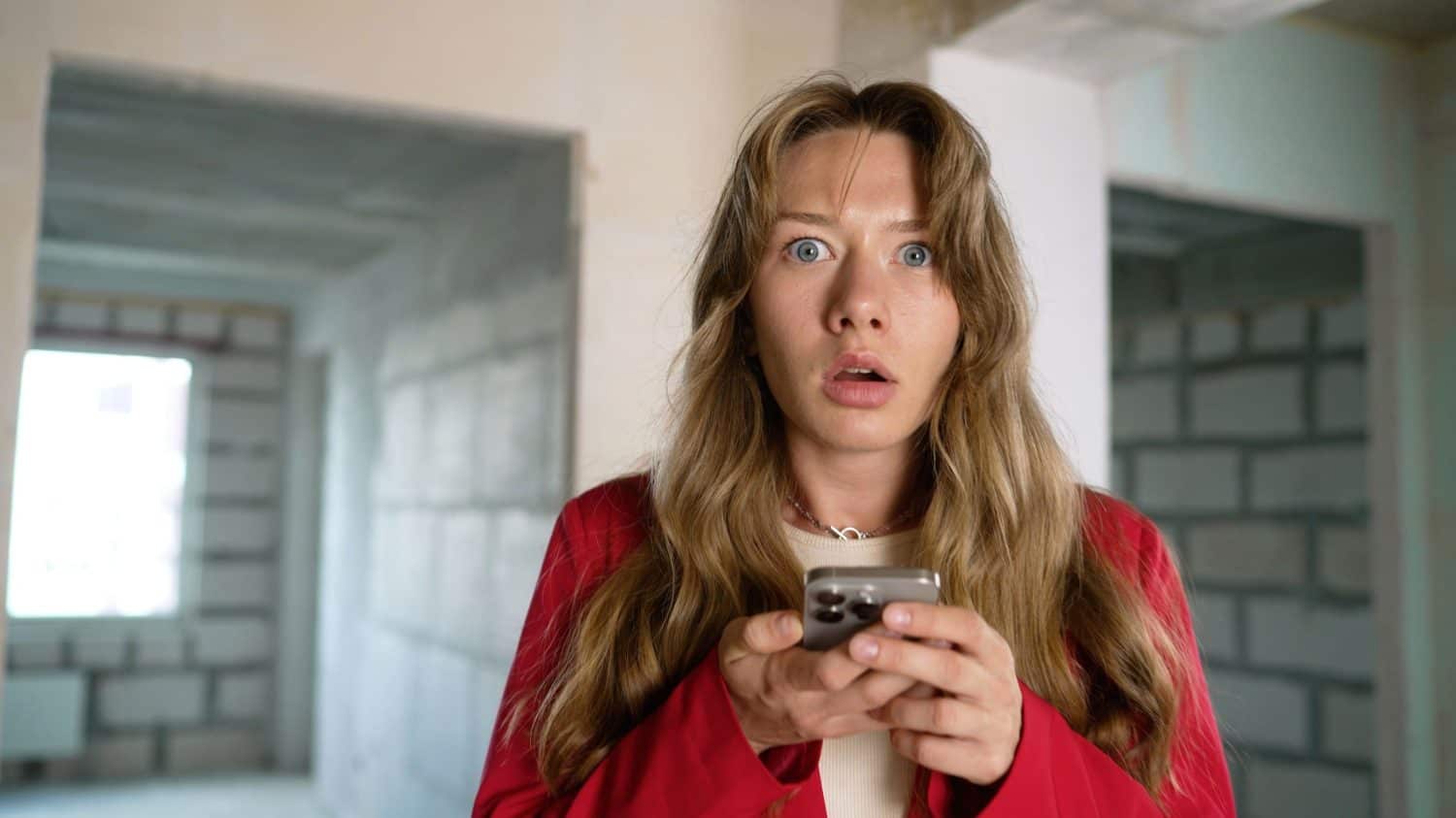 Shocked woman reading bad news on smartphone in unfinished apartment.