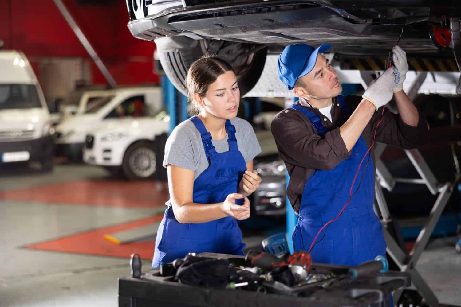 Proficient car mechanic checking operability of auto-element while girl in blue overalls watching him attentively in auto-repair shop