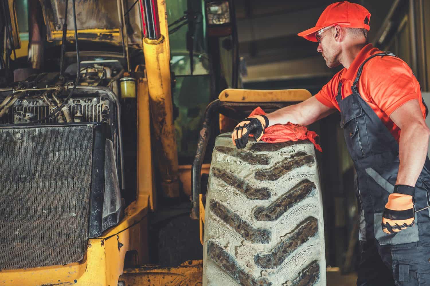 A mechanic inspects a large tire on a piece of construction machinery, focusing on repairs while wearing an orange cap and gloves in a well-lit workshop.