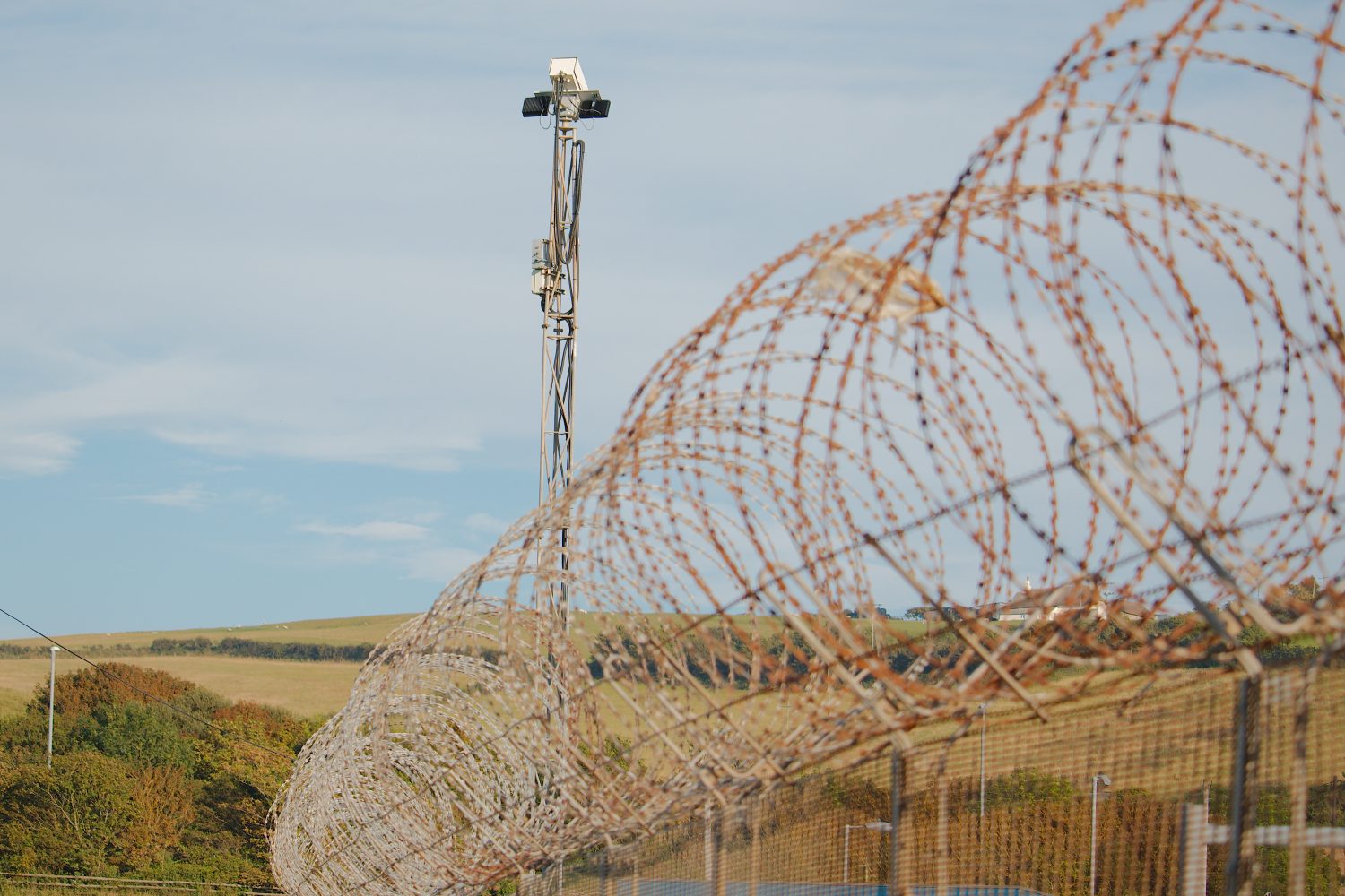 Razor wire fence with CCTV tower background protecting a secure facility.